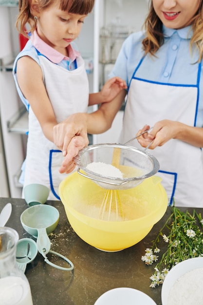 Young mother explaining daughter how to sift flour in big bowl when making pancake dough