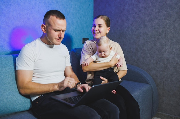 A young mother and father are sitting with their newborn son on the couch and watching a movie on a laptop Family relations with a small baby A family couple with a child at home on the sofa