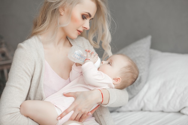 Young mother feeding her little cute baby daughter with bottle of child formula. Woman with her newborn baby at home. Mom taking care of a child. Alternative to breast feeding.