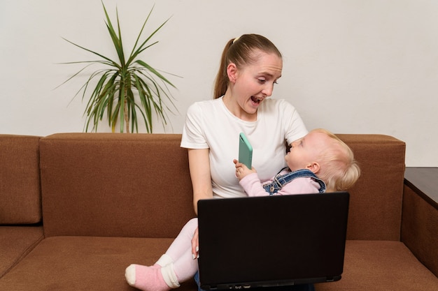 Young mother freelancer is playing with child and works in laptop.