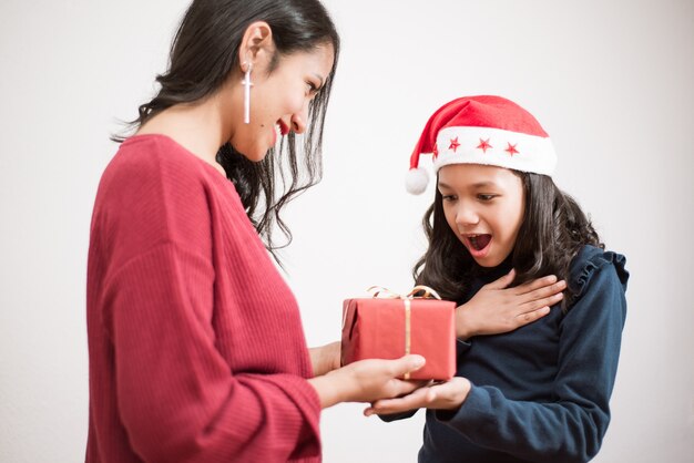 若い母親は、白い背景の上の娘にクリスマスのための赤いかわいいプレゼントを与えます。