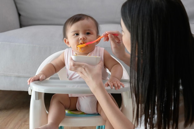 Giovane madre che aiuta il bambino a mangiare cibo misto sulla sedia del bambino