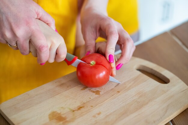 young mother helping daughter to cut tomato with kitchen knife on cutting board