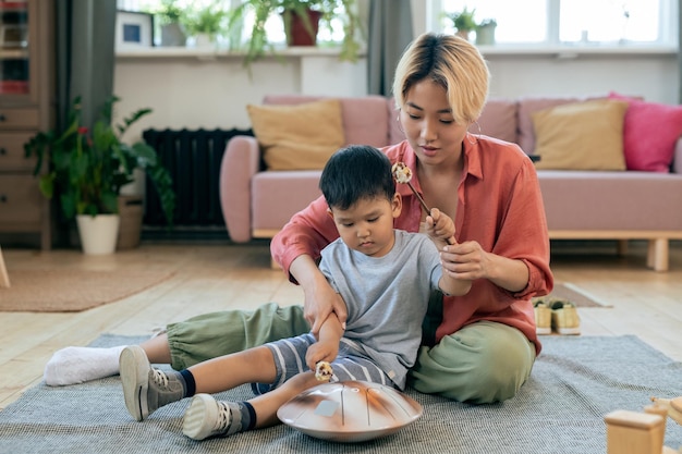 彼女のかわいい幼い息子がハングドラムを打つのを手伝っている若い母親