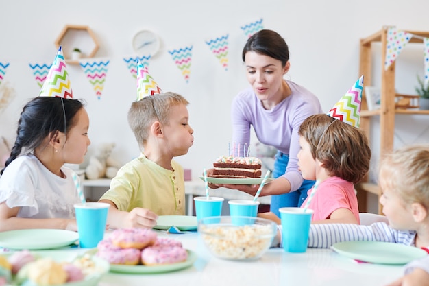 Young mother helping little kids to blow candles on birthday cake during home party around served table