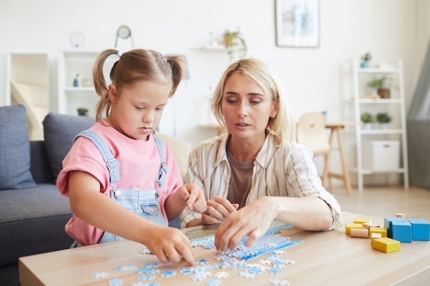 部屋のテーブルでパズルを収集するダウン症の娘を助ける若い母親