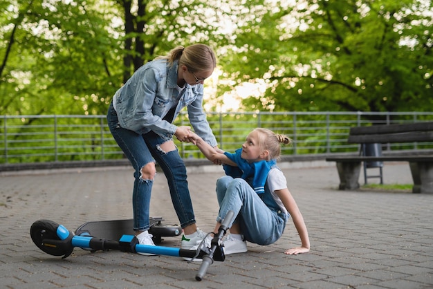 Young mother helps her daughter after falling accident e-scooter in city park