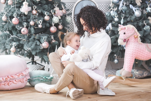 Young mother and her daughter having fun near the Christmas tree indoors