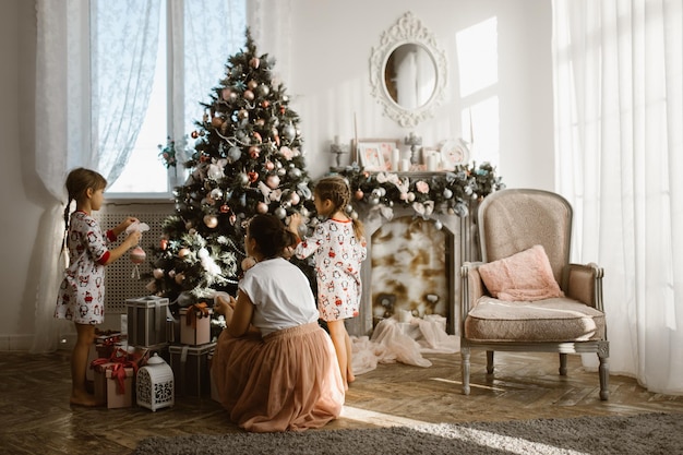 Молодая мать и ее маленькие дочери украшают новогоднюю елку в светлой уютной комнате