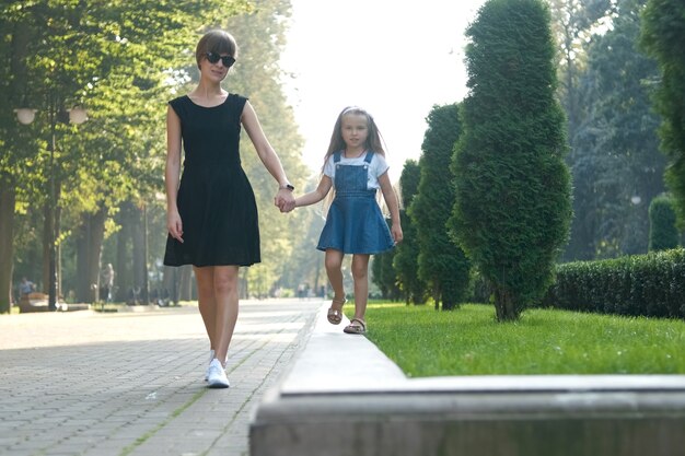 Young mother and her small daughter with long hair walking together holding hands in summer park.