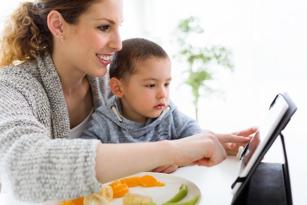 젊은 어머니와 집에서 과일을 먹는 동안 디지털 태블릿을 사용하는 그녀의 아들.