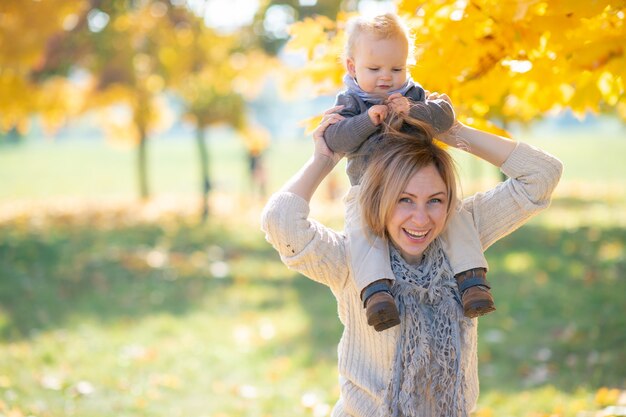 Young mother holding her  child on  shoulders in sunny park.