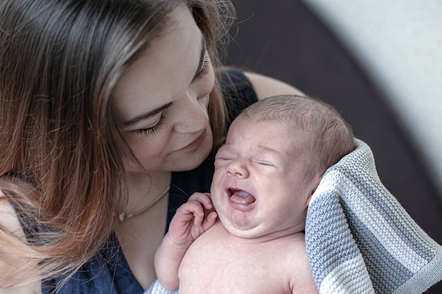 Young mother holds a crying newborn baby boy in her arms, close up.