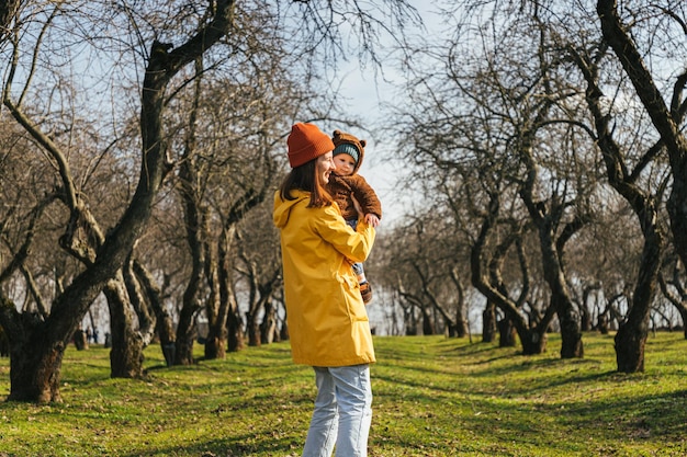 a young mother holds her child and looks at his hand in an alley of trees without leaves