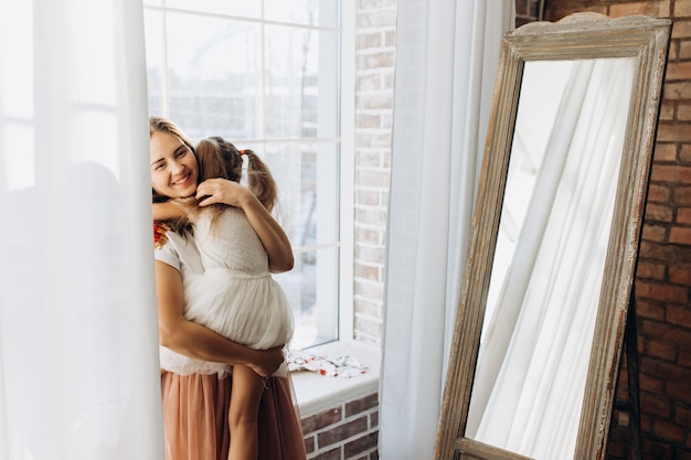 Young mother holds on her hands her little daughter next the window and the mirror in the light cozy room .