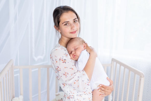 若い母親が生まれたばかりの赤ちゃんを腕に抱き、ベビーベッドの近くの寝室で母性と幸せな家族の概念を眠らせる