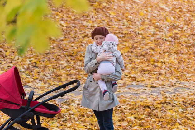 秋の公園で赤ちゃんを抱き締める若い母親。秋の森を歩く乳母車を持つ母。