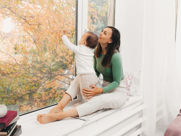 A young mother hugs her daughter, sitting in front of the window. Happy mom holding her baby while sitting on the windowsill