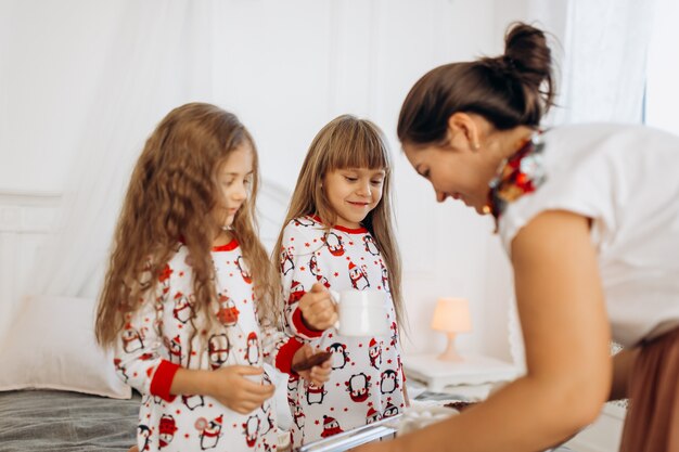 Young mother is bringing cocoa with Marshmallows and cookies to her daughters in pajamas sitting on the bed in the full of light cozy room .