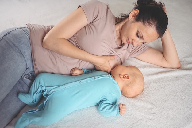 Young mother lies with a newborn cute infant baby in blue jumpsuit on the bed, hugging and breastfeeds him with breast milk. Healthy child, concept of hospital and happy motherhood. Nurser