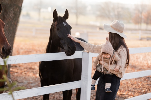 Young mother and little baby girl near a horses in autumn sunny day