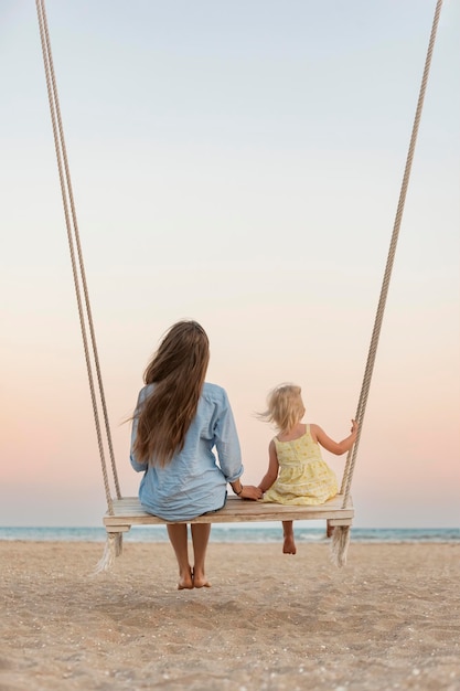 La giovane madre e la bambina bionda si siedono sull'altalena sulla spiaggia durante il tramonto mamma e figlia sulle magiche luci del tramonto del mare