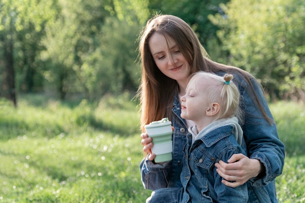 Young mother and little daughter against on green park background Mom drinks her daughter from silicone cup