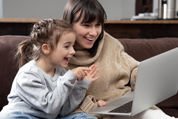 Young mother and little daughter with laptop computer at home sitting on sofa, feeling joy enjoying time together.