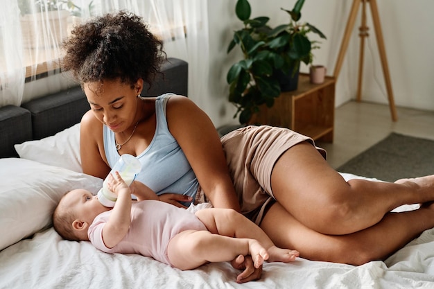 Photo young mother looking how her cute baby eating milk from bottle while they lying on bed