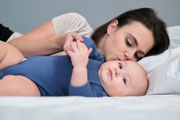 Young mother lovingly kisses her son toddler. Woman playing with baby while lying in bed