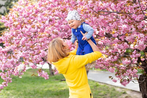 Young mother mom in medical mask holding her little baby son boy child under blossoming Sakura Cherry trees with falling pink petals and beautiful flowers