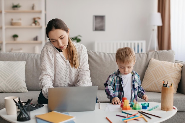 ラップトップを使用して携帯電話で話し、子供の息子の忙しいお母さんの世話をする若い母親のマルチタスク