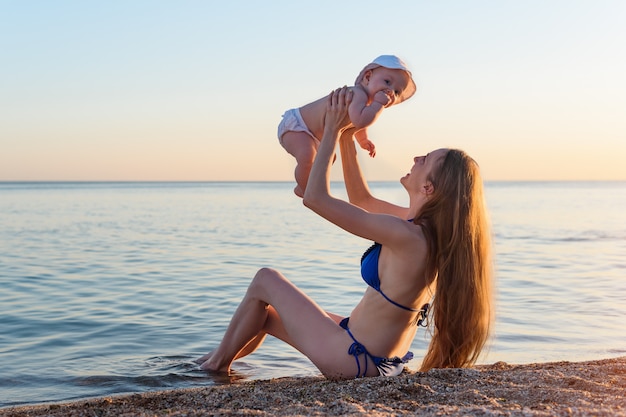 Young mother and newborn on beach on sea background. Mom catches baby. Vacation with child at sea