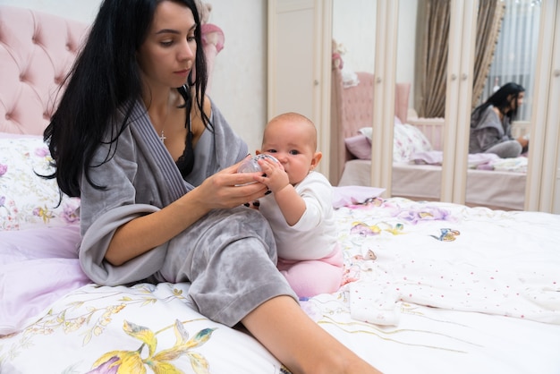 Young mother preparing a bottle feed for her little baby daughter as they relax together on a bed reflected in the mirror on the wardrobe