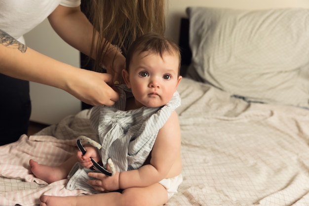 A young mother puts her baby in baby clothes. The baby obediently sits on the bed. Authentic lifestyle photos.