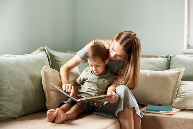 若い母親、彼女の子供に本を読んで、彼らの家のリビングルームで少年、窓から太陽の光