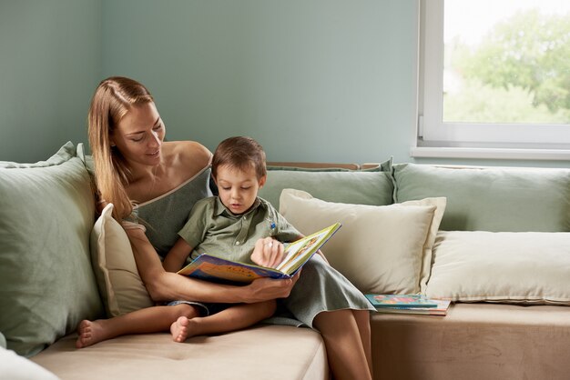 若い母親、彼女の子供に本を読んで、彼らの家のリビングルームで少年、窓から太陽の光