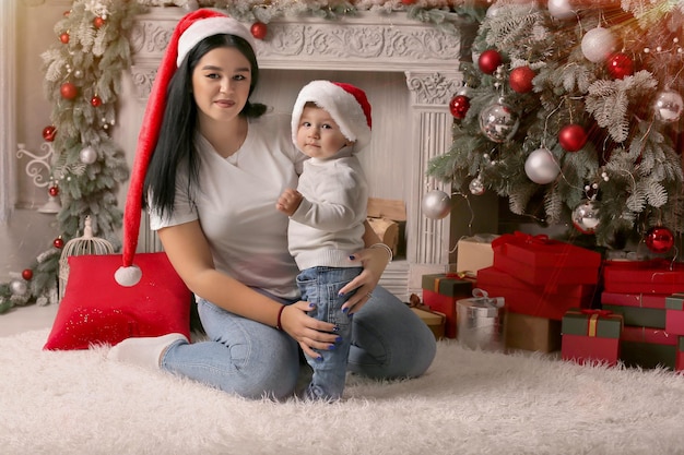 young mother in a Santa Claus hat hugs her little son by the fireplace and the Christmas tree