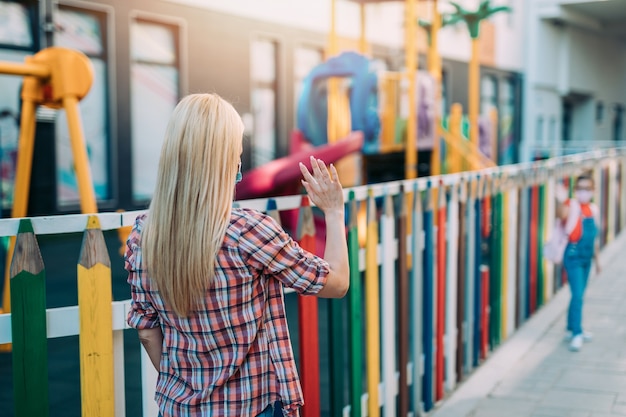 校庭で顔面保護マスクをつけた幼い娘に別れを告げる若い母親。彼らは顔面保護マスクを着用しています。学校のコンセプトに戻ります。