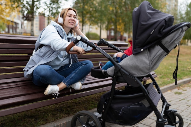 Молодая мама сидит на скамейке во время прогулки с коляской и слушает музыку в наушниках