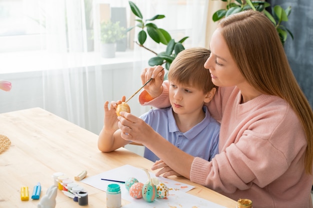 Young mother sitting at table with paint cans and adding paint on sons Easter egg while they making Easter decorations