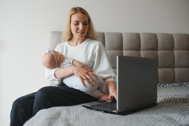 Young mother sitting with baby on bed and working at laptop from home