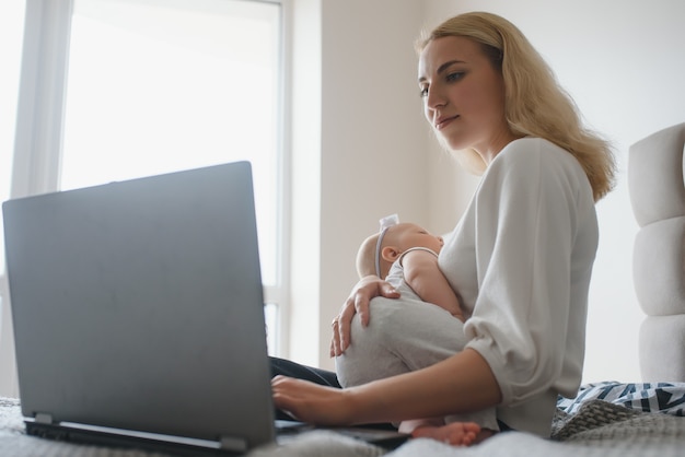 Young mother sitting with baby on bed and working at laptop from home