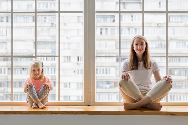 Young mother sitting with her 3-years daughter in lotus pose asana on window sill in front of window