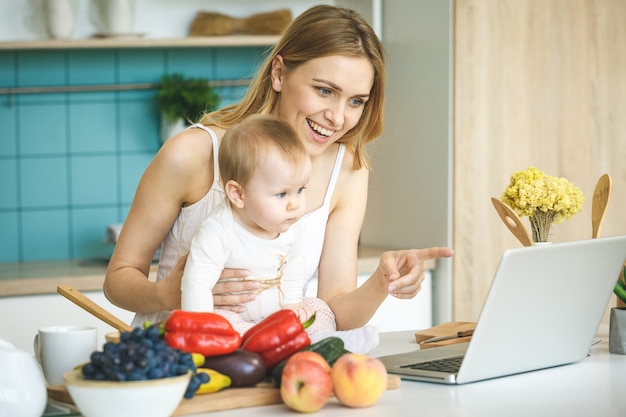 Young mother smiling, cooking and playing with her baby daughter in a modern kitchen. Using laptop.