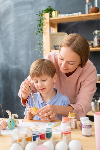 息子の後ろに立って、彼が卵にイースターのデザインを作るのを手伝っている若い母親