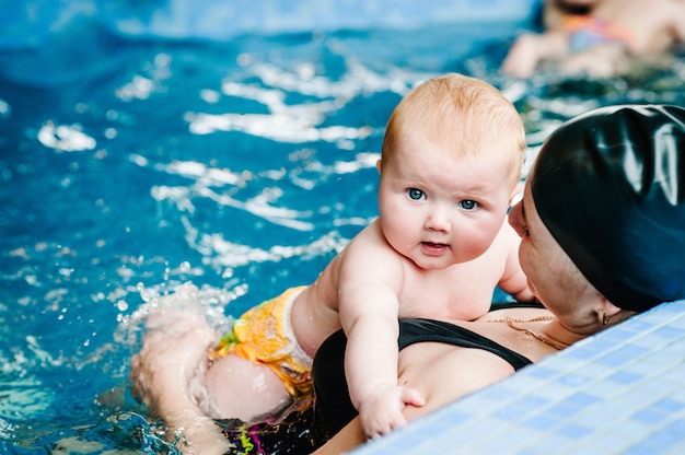 Young mother, swimming instructor and happy little girl in paddling pool. Teaches infant child to swim. Enjoy. Mom holds hand child preparing for diving. doing exercises. baby lying on the mother body