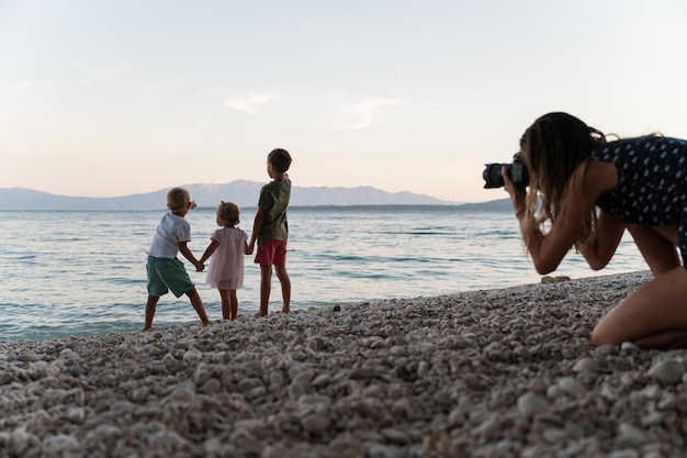 Photo young mother taking photos of her three kids standing on pebble beach looking in the distance, with professional dslr camera.