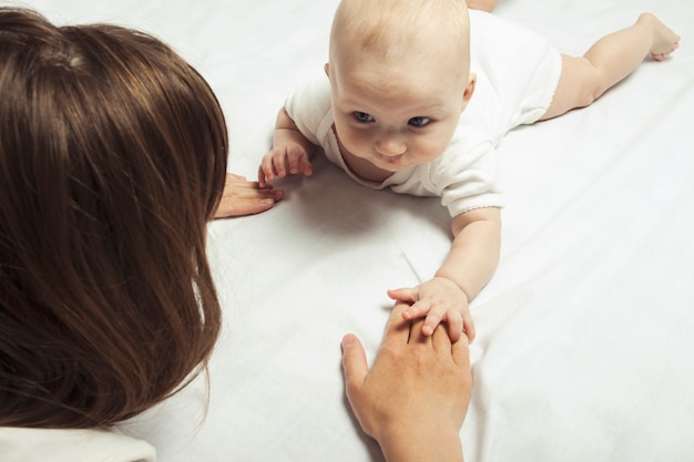 Young mother teaches to crawl a little baby on the bed with a white sheet