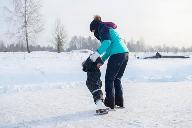 若い母親が赤ちゃんの息子にアウトドアスケート場でアイススケートを教えています 家族はアイススケート場で冬を楽しんでいます